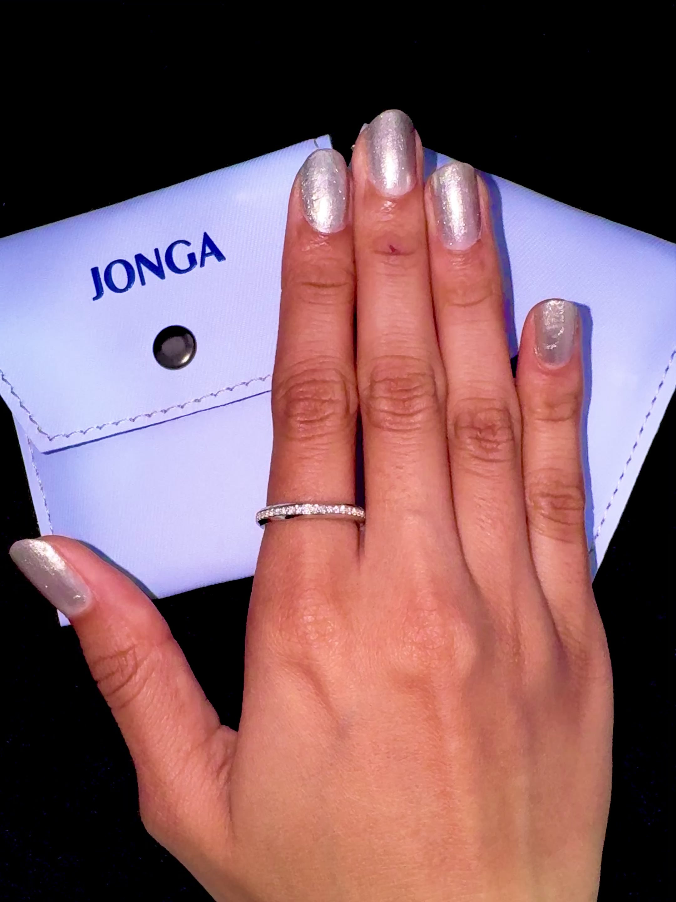 Elegant depiction of a white gold paved band adorning a finger in a lifestyle “ring on her” shot. The ring highlights a dazzling central design featuring pavé, full pave, scattered pave, and cluster pave techniques, set in luminous white gold—also referred to as “opaline” or “milky gold.” The surrounding glossy finish adds a touch of modern allure and refined sophistication. Keywords: graceful, intricate, luxurious, chic, contemporary design.
